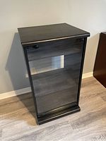 Photo of a black or dark wood record player cabinet with a clear glass front door, showing interior shelving and simple rectangular design.