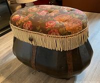Closed view of the storage chest showing floral upholstered cushioned lid with beige tassel fringe trim over dark rounded wooden base with embossed strips.