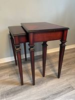 Two dark wood nesting side tables arranged stacked one under the other on a wooden floor against a grey wall.