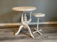 Photo of two white wooden side tables, one large round top with carved spindle leg and three curved feet, and a smaller hexagonal top with turned spindle leg and four-foot base, placed on wood floor against beige wall.