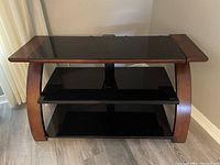 Photo shows a TV stand with a dark wooden frame and two black glass shelves, placed on light grey flooring near a white curtain.