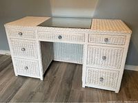 White wicker desk or dresser with six drawers and glass top inset against a gray wall on wood floor.