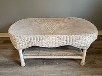 White wicker oval coffee table without glass, with woven top and lower shelf, four legs, placed on wooden floor.