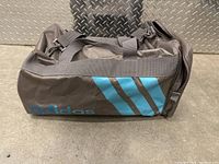 Full side view of Adidas gym bag showing blue stripes and logo on gray fabric, lying on diamond plate metal surface.