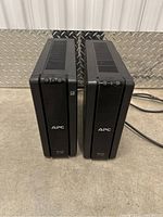Two black APC Back-UPS XS 1300 units standing side by side on a concrete floor against a metal wall