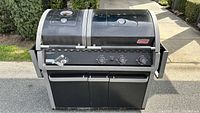Front view of Coleman Evenheat gas BBQ grill showing control panel with three knobs, temperature gauge on lid, double lids, and storage cabinet below