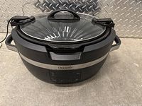 Black Crock-Pot slow cooker with clear glass lid and handles, showing overall unit from front angle on concrete floor.