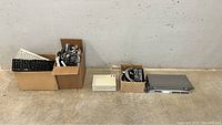Photo of the entire lot showing 4 cardboard boxes with keyboards, cables, a KVM switch, and a server/network device on a concrete floor