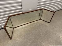 Tri-fold mirror showing three connected rectangular panels framed in brown wood, positioned on concrete floor near a white industrial wall.