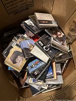 Photo of numerous music CDs in jewel cases piled in a cardboard box. Album covers with artist photos and artwork visible.
