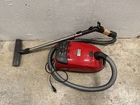 Red Miele Cat & Dog Plus vacuum on concrete floor showing hose, metal wand, and floor head with power cord attached