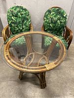 Front view of two rattan armchairs with green tropical leaf print cushions and round rattan table with glass top in between.