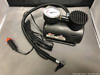 Auto Air Compressor Model COM-250 with pressure gauge, hose, and power cord with 12V plug, shown outside packaging on a reflective surface.
