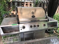 Front view of Vermont Castings stainless steel propane BBQ grill showing four control knobs, temperature gauge on hood, left and right side shelves, and under cabinet storage doors.