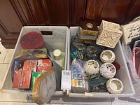 Two white plastic bins filled with candles, incense boxes, decorative candle holders, and accessories, shown from above