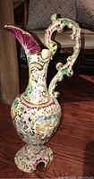 Full view of ornate porcelain vase pitcher with curved handle and floral decorations on wood floor