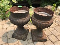 Pair of two cast iron planters placed outdoors on stone tile patio. Show full view of planters from above, displaying weathered and rusty interiors.