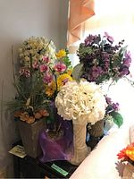 Grouped photo showing multiple faux flower arrangements including white hydrangea-like flowers, pink orchids, purple berries, and yellow daisies placed on and around beige and ceramic vases.