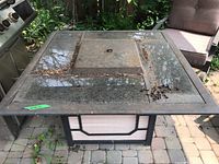 Square outdoor fire table with black metal frame and tinted glass top panels. Center has raised square metal section with metal ring. Shows dirt, leaves, rust, and screws on top.