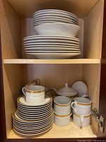 Photo showing stacked dinner plates and soup bowls on top shelf, teacups, saucers, mugs, teapot, and sugar bowl on bottom shelf inside a wooden cupboard.