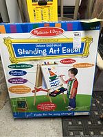 Front view of boxed Melissa & Doug Deluxe Solid-Wood Standing Art Easel showing product features and child using easel.