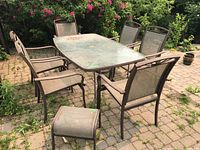 Front view of rectangular glass top metal patio table with six mesh armchairs and one ottoman around it, situated on a brick patio with greenery in the background.