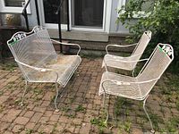 Photo of white painted cast metal settee and two chairs with mesh seating and backs outdoors on paved surface showing rust and wear