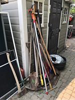 Wide view of garden tools leaning against shed including rakes, shovels, broom, lopper, and garden bed edging materials.