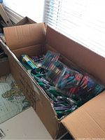 Large open cardboard box filled with packs of pens and loose pens and pencils on a table.