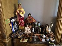 Overview of religious statues, crosses, prayer candles, and small figurines placed on an old wooden dresser.