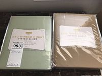 Two king size fitted sheets sealed in clear plastic packaging. One is sage green, the other beige, both labeled 250 thread count, 100% cotton from brand home Q.