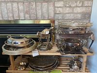 Full view of assorted silver plated serving dishes on wooden rack with various trays, warming trays, plates, and a gravy boat.