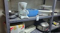 Full shelf view showing Rival automatic steamer, blue mixing bowls, rice cooker, coffee maker, and supply box below