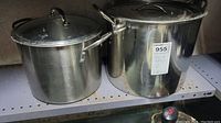 Two aluminum stock pots on a metal shelf, showing front and side views, both with lids and handles.