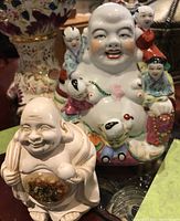 Photo of two Buddha figurines, larger porcelain with colorful small figures and smaller cream-colored with painted belly design.