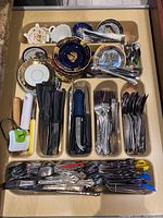 Top view of drawer containing assorted kitchen utensils including knives, forks, spoons, and decorative china plates and saucers.