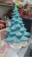 Full view of bluish-green ceramic Christmas tree sitting on curved white base with light holes visible on branches.