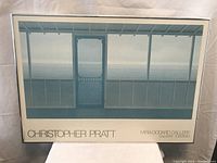 Photo showing framed blue water artwork by Christopher Pratt titled "Cottage" with a glass front, measuring about 2 feet by 34.5 inches.
