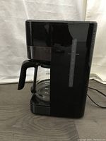 Black Oster coffee maker from side, showing glass carafe and water level indicator.
