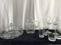 Full set of glassware including small glasses, medium glasses, wine glasses, water jug, and cheese & cracker plate with lid placed on dark cloth background against white draped backdrop.