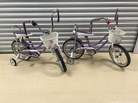 Two matching purple Schwinn girl child bikes with white baskets and training wheels seen from front angle
