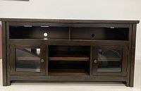 Front view of the wooden TV stand showing two glass door cabinets on the sides, open middle shelves, and top compartments with cable holes.