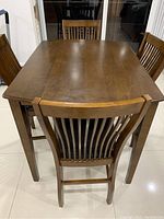 Photo showing rectangular wooden dining table with four matching wooden chairs, view from one short side. Table surface smooth with slight wear visible. Chairs have vertical slat backs and matching medium brown finish.