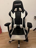 Front view of black and white S-RACER gaming chair with prominent headrest logo, padded armrests, and caster wheels on floor.