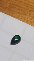 Pear-shaped black opal cabochon gemstone resting on a white surface to highlight color and shape.