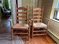 Photo showing two wooden arm chairs with woven rush seats and ladder backs placed side by side on a wooden floor near a window. Chairs have carved detail on the top horizontal slat and vertical posts with finials.