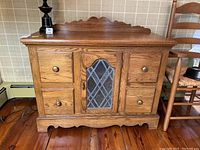 Front view of wooden cabinet showing four drawers and decorative glass door with diamond leaded glass panel.