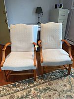 Front view of two matching upholstered arm chairs with wooden frames and beige fabric upholstery on seat and backrest.