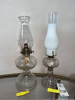 Two glass hurricane oil lamps side by side on a mirrored surface. One lamp has clear glass chimney and base, the other has frosted white glass chimney with textured clear glass base.