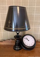 Photo of black lamp with paper shade and black plastic base with gold decorative accents and black round clock with white face and black frame.